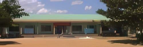 A single-story building with a green roof and a red sign above the entrance. Trees partially frame the structure, and the sky overhead is partly cloudy.