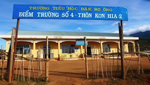 A yellow school building with a blue sign in Vietnamese, reading "Trưá»ng Tiểu Há»c Äăk RÆ¡ Ông, Äiểm Trưá»ng Số 4 - Thôn Kon Hia 2," stands behind a fence on a dirt yard under a clear sky.