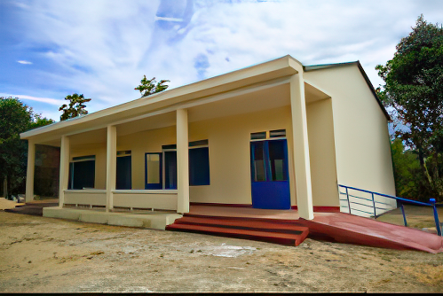 Single-story building with beige walls, blue doors and windows, and a red entry ramp surrounded by a sparse, natural setting and partly cloudy sky.