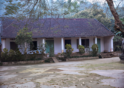 A single-story building with a sloped roof, surrounded by trees and greenery. It has multiple pillars supporting a front porch and a concrete courtyard in front.