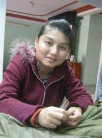 A person wearing a maroon hooded jacket with fur trim is sitting indoors, leaning forward slightly, and looking at the camera.